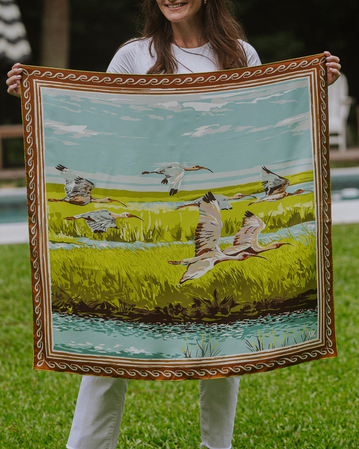 Woman holding a silk scarf with birds flying over a marsh and a decorative border.