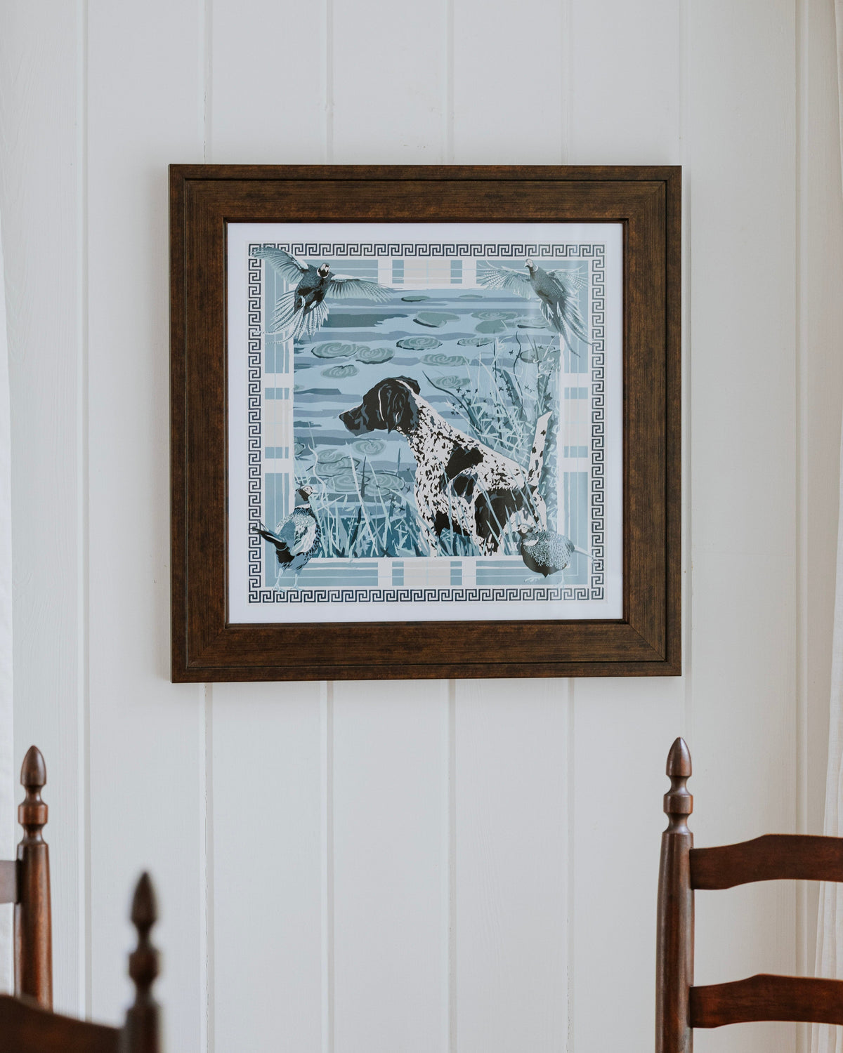 A framed square art print featuring a dog in a blue-toned hunting scene, hung on a white paneled wall above a wooden dining table and chairs.