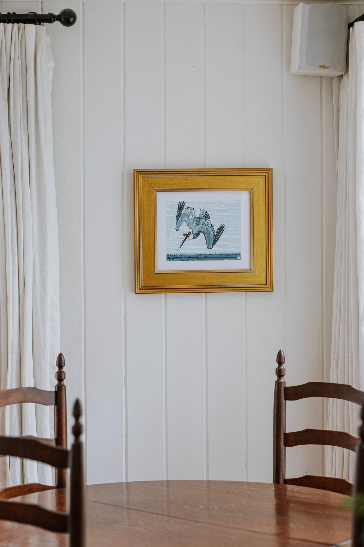 A framed art print depicting a diving pelican in a gold frame, hanging on a white paneled wall above a wooden dining table and chairs.