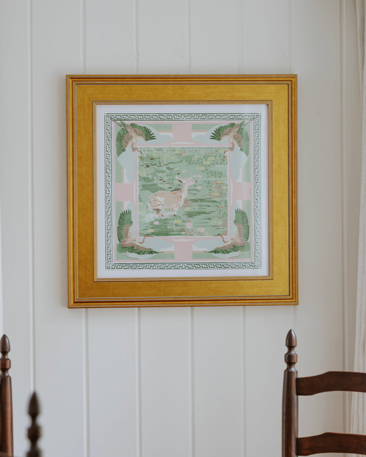 A framed square art print with a gold frame hangs on a white paneled wall above a wooden dining table and chairs.