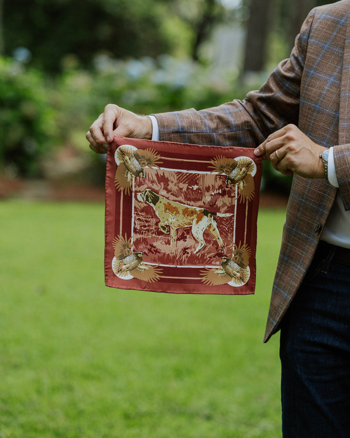 A person outdoors holding up a silk pocket square with hunting dog illustration and decorative corner details.