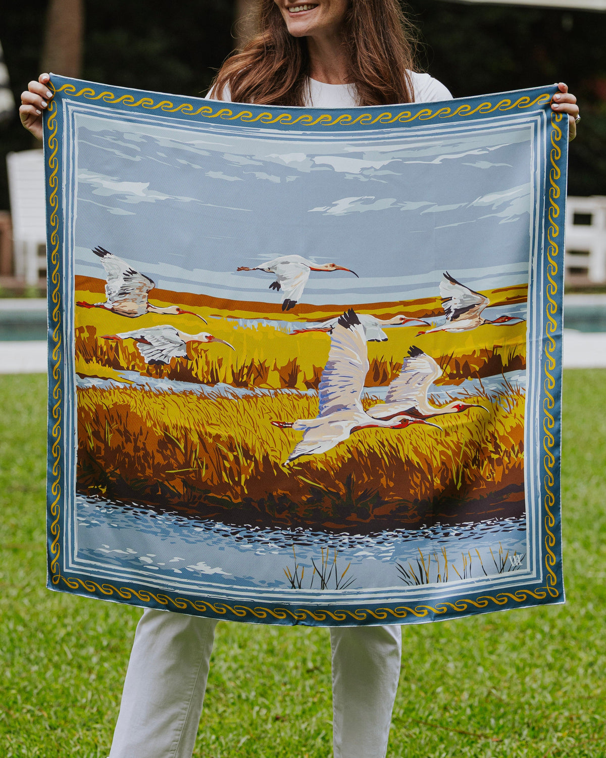 A woman holding up a square silk scarf  featuring a painted landscape with white birds over golden marsh grass under a blue sky. 