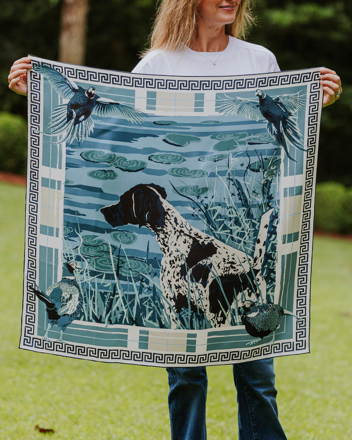 A woman a square silk scarf featuring a blue and white illustration of a black-and-white dog in a marsh with birds and a Greek key border.