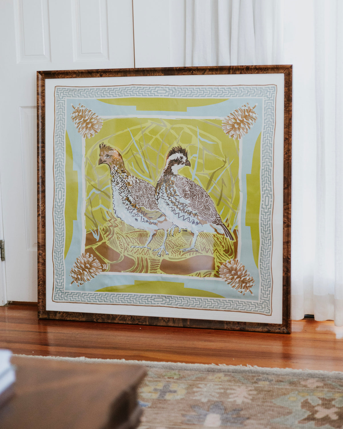 A framed silk scarf featuring two bobwhite quail on a yellow and green background, leaning against a white paneled wall on a wooden floor.