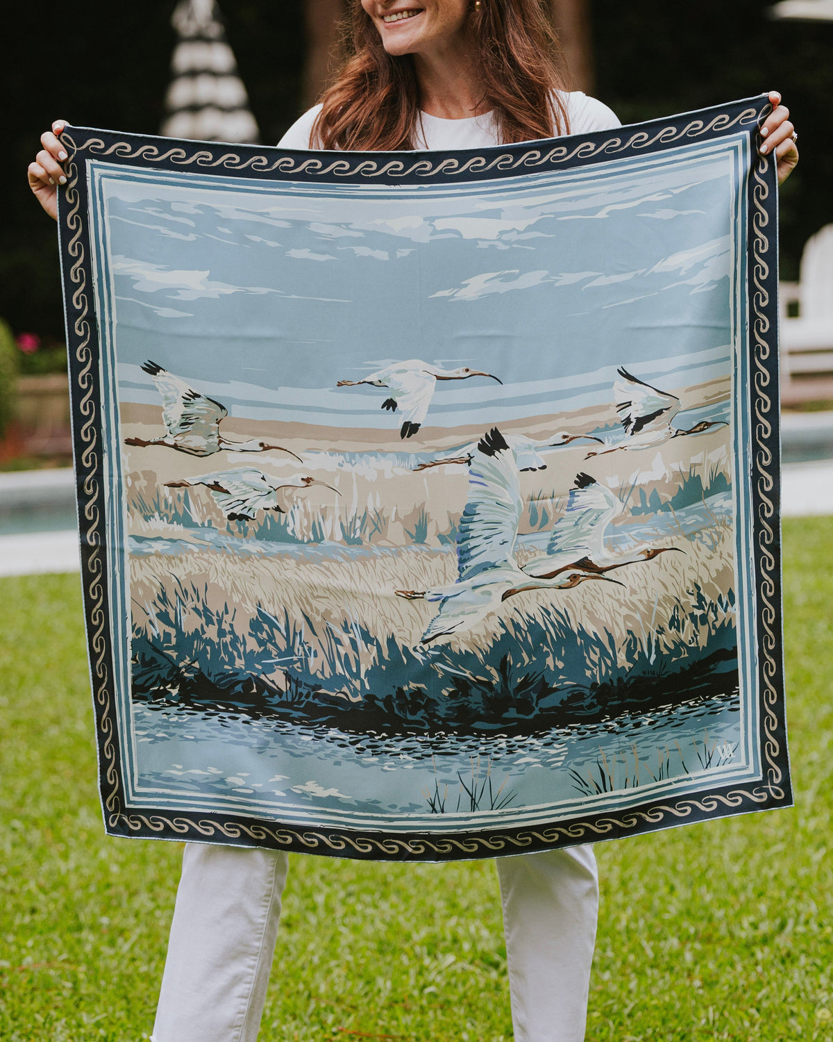 Woman holding a silk scarf with birds flying over a marsh and a decorative border. 
