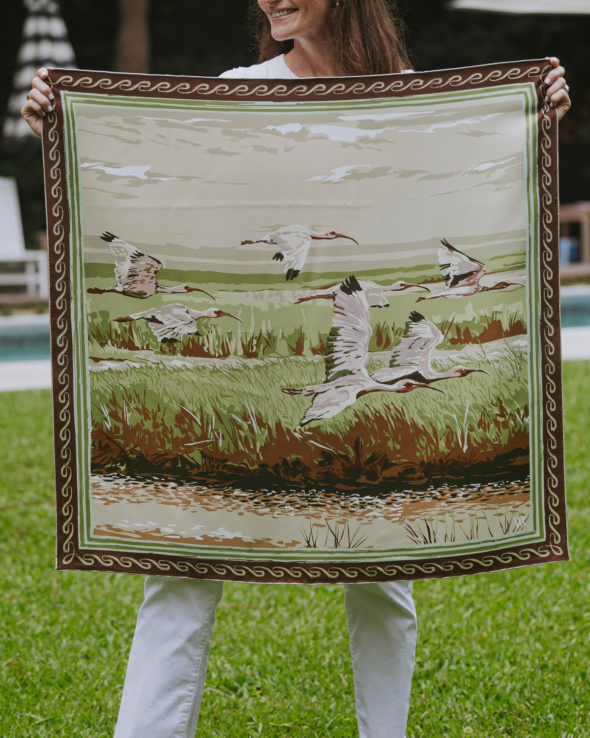 A woman holding up a large square silk scarf featuring a marshland scene with birds. 