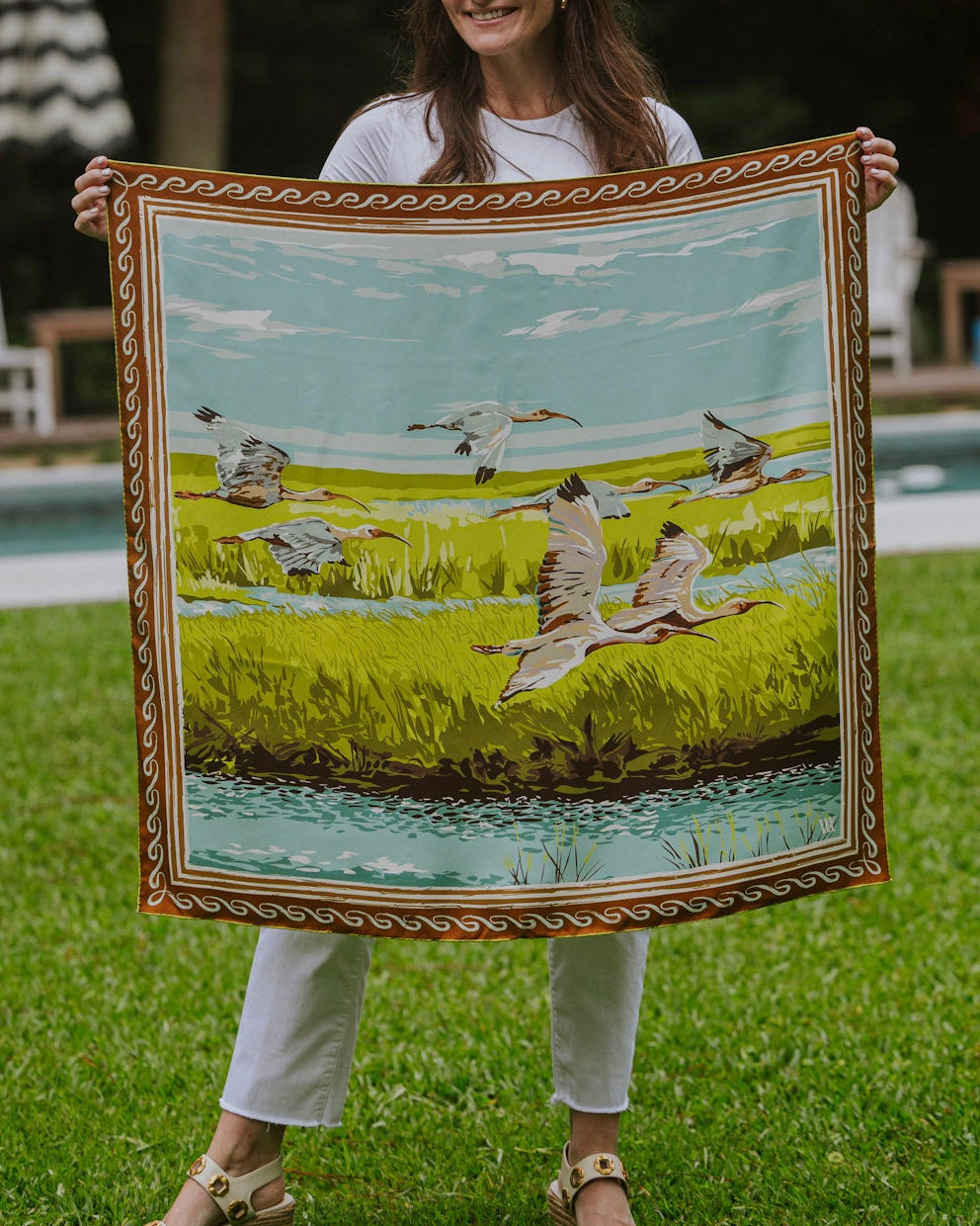 A woman stands outdoors holding up a large silk scarf featuring a lush green marsh with birds and a light blue sky.