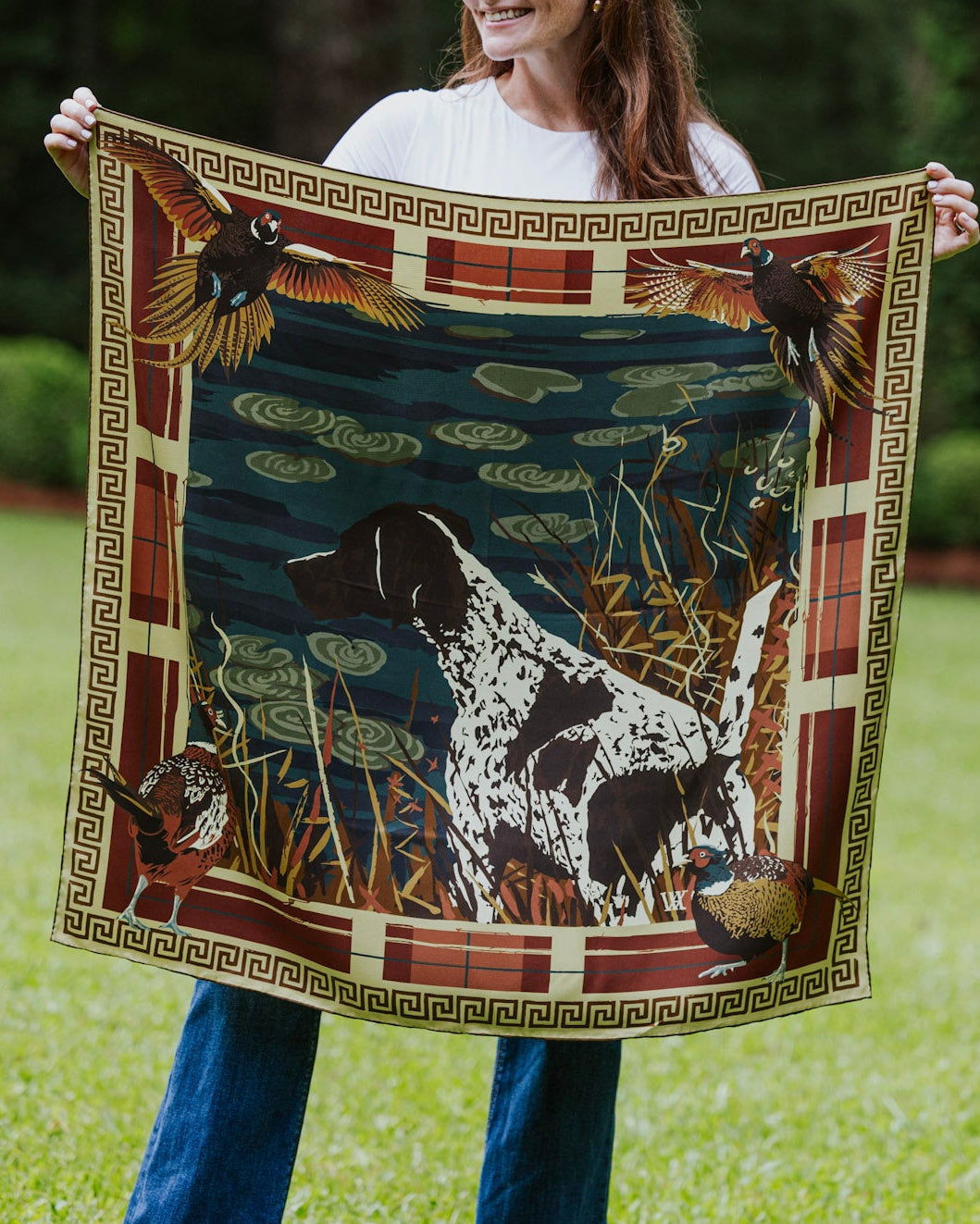A woman standing outdoors holding up a silk patterned scarf. The scarf features a dark blue pond scene with a black-and-white dog, ducks, lily pads, and a decorative border.
