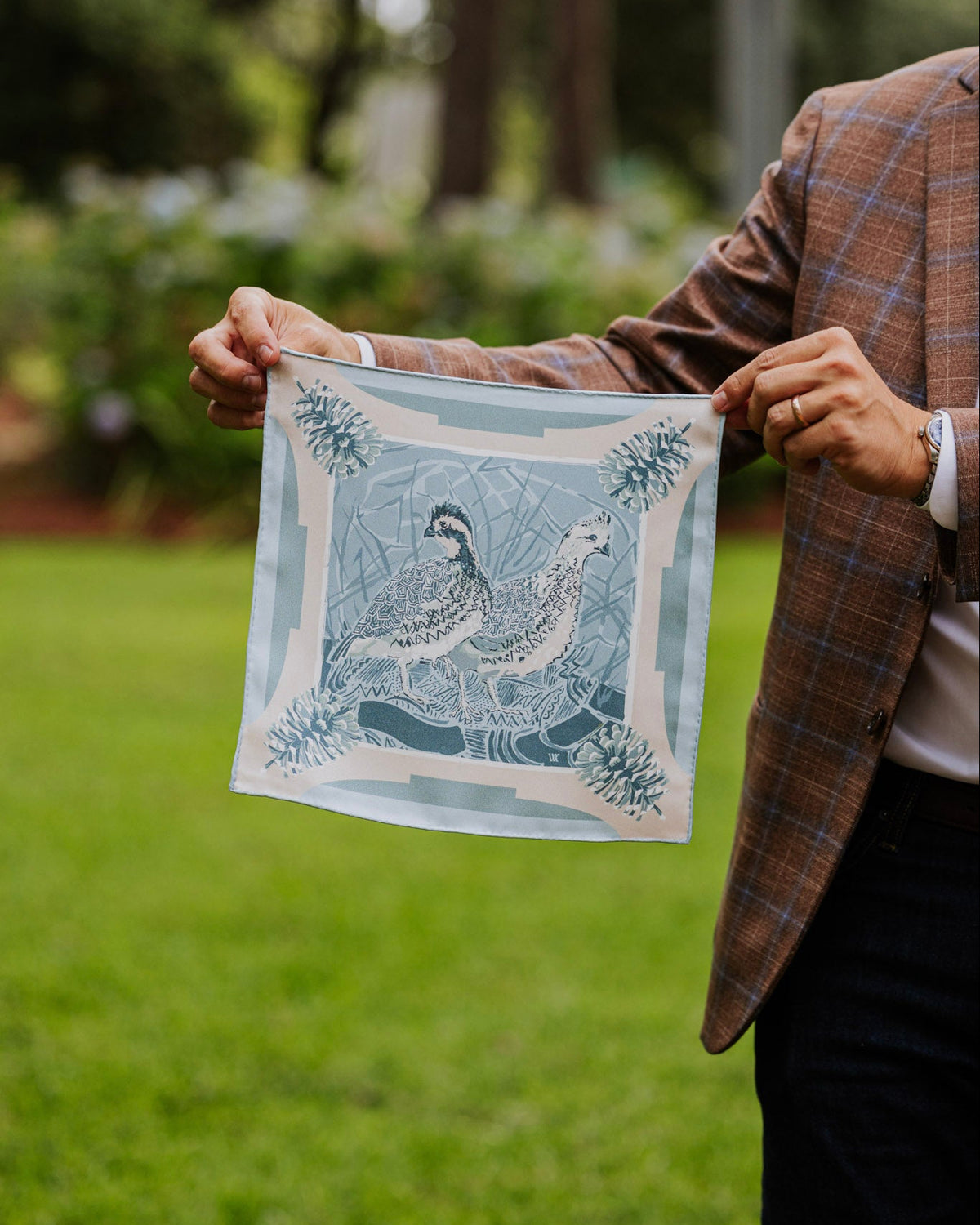 Man holding a silk pocket square in a monochromatic blue color featuring quail and pinecones. 